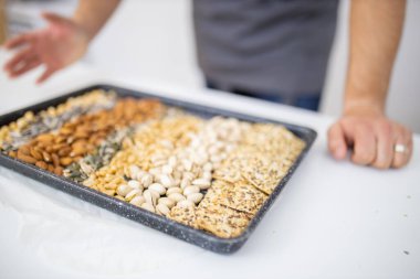 Siyah bir tepsi fındık ve tohumla beyaz bir masada oturan adam.