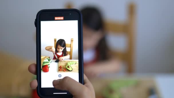 Vrouwelijke handen met een smartphone opnemen van een klein meisje snijden van een avocado — Stockvideo