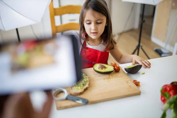 Mão com um smartphone gravando uma menina cortando um abacate — Fotografia de Stock