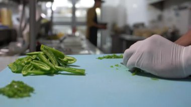 Eldivenli eller mavi kesme tahtasındaki biber dilimlerini yavaşça kesiyor.