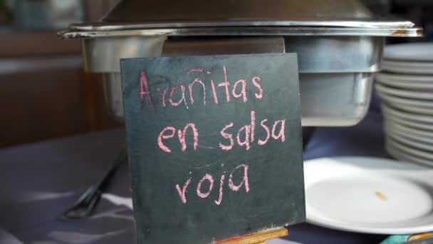 Μεγάλο μεταλλικό δοχείο και μαύρο σημάδι με ροζ ισπανικό κείμενο σε ένα τραπέζι — Αρχείο Βίντεο