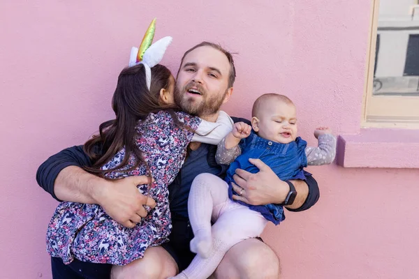 Happy father holding his two adorable daughters with pink wall as background
