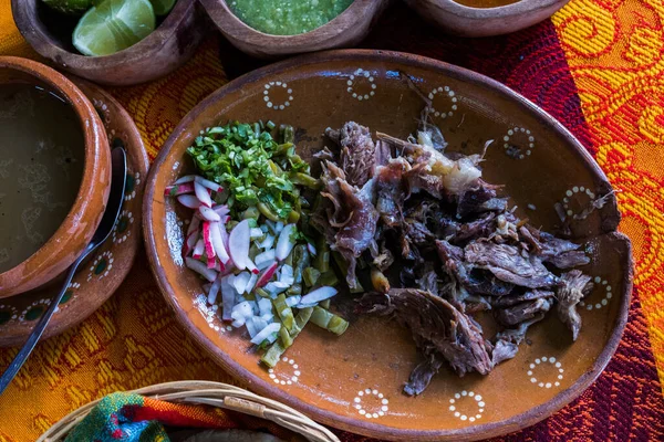 Mexican chopped lamb meat, hot sauces, and broth on colorful tablecloth