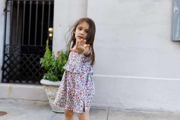 Красивая маленькая девочка в цветущем платье позирует перед серой стеной — стоковое фото