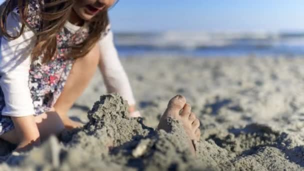 Adorable little girl covering adult feet with sand at the beach — Stock Video