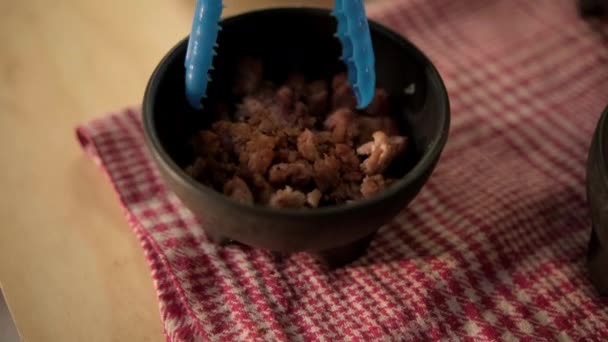 Niebieskie szczypce chwytające siekaną wieprzowinę z czarnej miski — Wideo stockowe