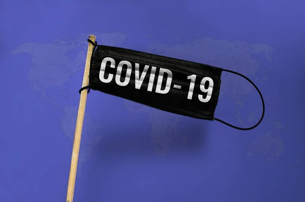 Tıbbi maske siyah bayrak direğin üzerinde COVID ile dalgalanıyor. Mavinin üzerindeki siyah maske dünya haritası dokulu arka plan. Virüs saldırısı bayrağı. Coronavirus COVID-19 kavramı