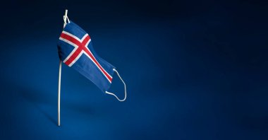 Koyu mavi arka planda İzlanda maskesi. Direğe tıbbi maske takmış İzlanda bayrağı sallıyordu. Virüs saldırısı bayrağı. Salgın koronavirüs COVID-19 'a karşı mücadele bayrağı kavramı. Boşluğu kopyala