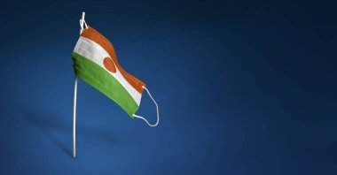 Koyu mavi arka planda Nijer maskesi. Direkte tıbbi maskenin üzerine Nijer bayrağı sallıyordu. Virüs saldırısı bayrağı. Salgın koronavirüs COVID-19 'a karşı mücadele bayrağı kavramı. Boşluğu kopyala