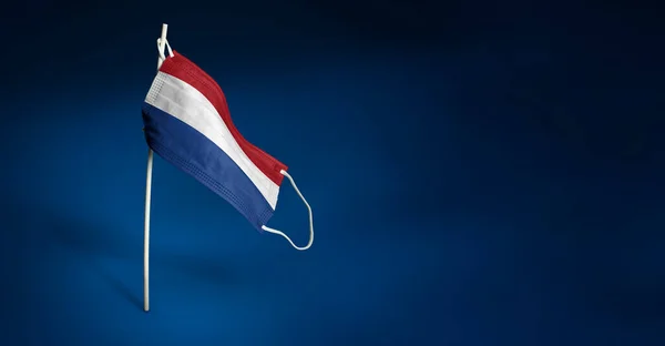 Nederlands masker op donkerblauwe achtergrond. Wuivende vlag van Nederland geschilderd op medisch masker op paal. Concept van het spandoek van de strijd tegen het epidemische coronavirus COVID-19 — Stockfoto