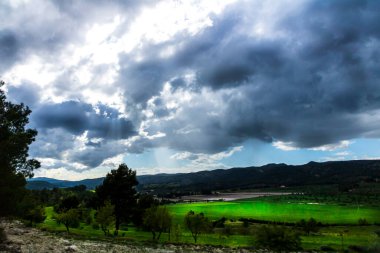 Bir günde yeşil çayır, beyaz ve gri bulutlar ve arkasında dağlar olan güneş ışınları..