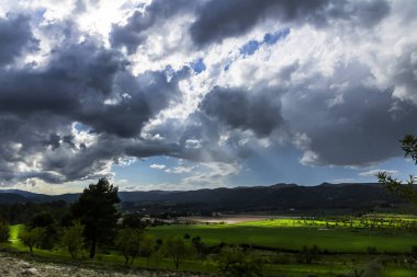 Bir günde yeşil çayır, beyaz ve gri bulutlar ve arkasında dağlar olan güneş ışınları..