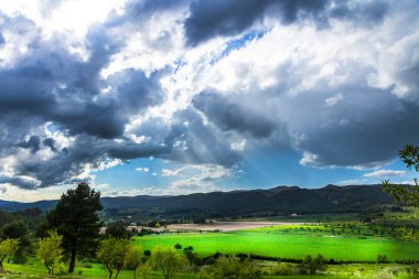 Bir günde yeşil çayır, beyaz ve gri bulutlar ve arkasında dağlar olan güneş ışınları..