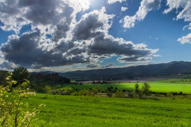 Bir günde yeşil çayır, beyaz ve gri bulutlar ve arkasında dağlar olan güneş ışınları..