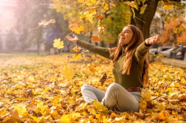 Parkta oturan gülümseyen kız ağaçların sarı yapraklarını fırlatıyor. sonbahar sezonu
