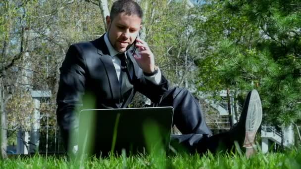 Empresario trabaja con portátil y teléfono móvil en el parque de la ciudad — Vídeos de Stock