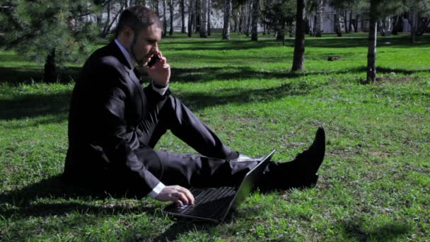 Biznesmen współpracuje z laptopa i telefon komórkowy w parku miejskim — Wideo stockowe