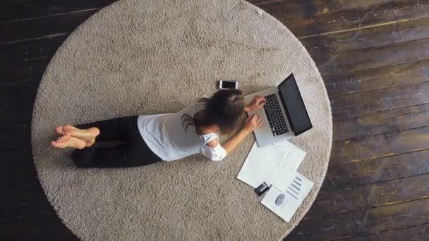 Молода брюнетка лежить на килимі і використовує ноутбук — стокове відео