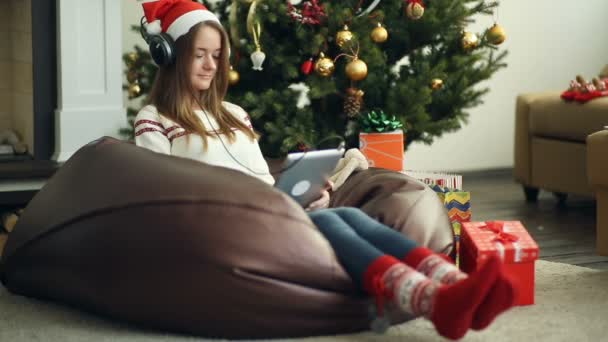 Chica sonriente en gorra de Navidad roja y calcetines usando almohadilla contra el árbol de Navidad — Vídeos de Stock