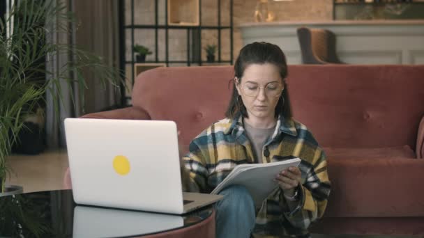 Mujer seria del milenio tomando notas en el cuaderno mientras se utiliza el ordenador portátil — Vídeos de Stock