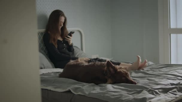 Vrouw gebruik haar mobiele telefoon op bed met haar hond — Stockvideo