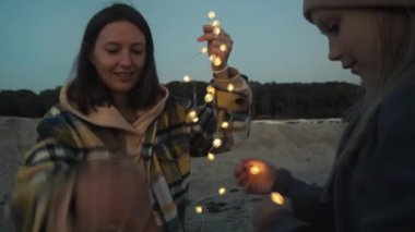 Genç bir kadın ve kızı sahilde ışıklarla oturuyorlar.