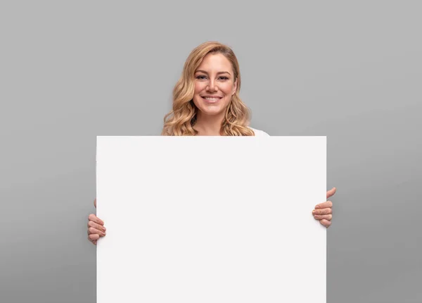 Mladá žena drží velký bílý prázdný list na šedém pozadí — Stock fotografie