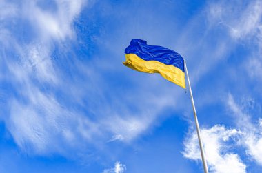 Büyük bir Ukrayna bayrağı, parlak mavi ve sarı yatay şeritleriyle, dramatik, parlak mavi gökyüzüne karşı dalgalanıyor, ince beyaz bulutlarla dolu, alçak bir açıdan bakıldığında.