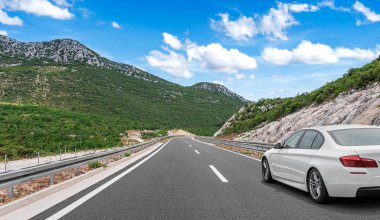 Beyaz bir araba, güneşli bir günde, yemyeşil tepeler ve kayalık yamaçlarla çevrili, açık mavi bir gökyüzünün altında, boş bir dağ yolunda ilerliyor..