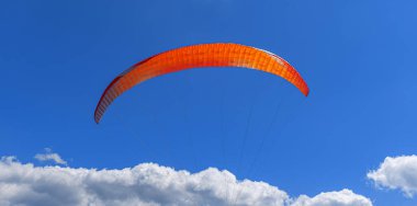 Aşağıdaki tüylü beyaz kümülüs bulutları tarafından çerçevelenmiş, derin mavi gökyüzüne doğru yükselen canlı bir turuncu ve gümüş paraglider kanadı özgürlüğü ve macerayı simgeliyor..