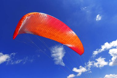 Yoğun bir turuncu ve kırmızı paraglider kanadı, parlak, güneşli bir günde, tüylü beyaz kümülüs bulutlarıyla kaplı koyu mavi gökyüzüne doğru yükselir..