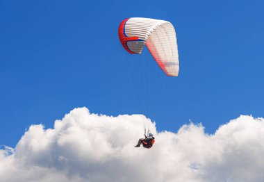 Bir paraglider açık, derin mavi gökyüzünde asılı kalır, güneşli bir açık hava macerası sırasında beyaz, tüylü bulutların üzerinde yavaşça süzülür..