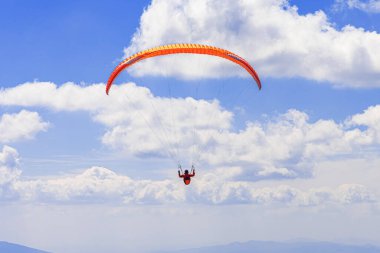 Turuncu gölgelikli bir paraglider, uzak dağların üzerinde yükselen beyaz kümülüs bulutlarıyla dolu parlak mavi bir gökyüzüne karşı siluetlendi..