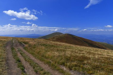 Çimenli, yuvarlanan bir dağ arazisinde savrulan toprak bir yol. Dağılmış bulutlarla mavi bir gökyüzünün altında..