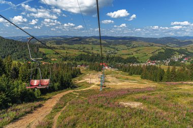 Bir teleferik pembe kır çiçekleriyle bezenmiş çimenli bir kayak yamacına iniyor, yemyeşil kaygan tepeler, dağınık ormanlar ve parlak mavi bir gökyüzünün altındaki küçük bir köy panoramik bir manzara sunuyor.