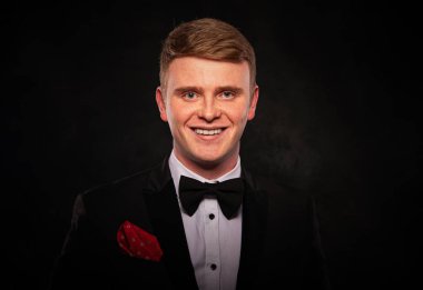 Siyah smokin ve papyon giymiş, koyu dokulu arka planda gülümseyen, kızıl saçlı bir gencin stüdyo portresi..