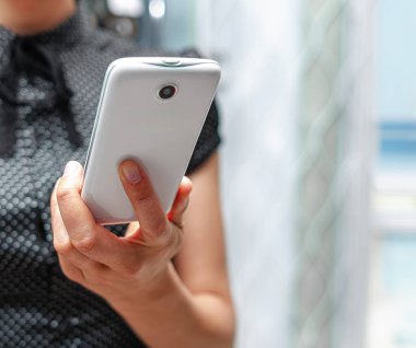 Siyah desenli gömlekli bir kişi beyaz bir akıllı telefonu pencerenin yanında tutuyor..