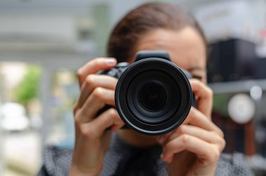 Siyah bir DSLR kamera tutan bir kişi, açık hava ortamında büyük merceği doğrudan izleyiciye odaklıyor..