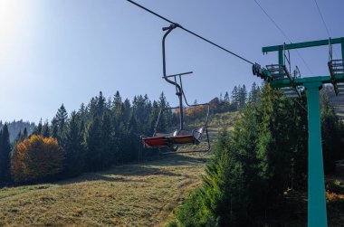 Açık mavi gökyüzünün altında yeşil ve yaprak döken ağaçlarla çevrili çimenli bir dağ yamacının üzerinde asılı duran bir kayak asansörü sandalyesi..