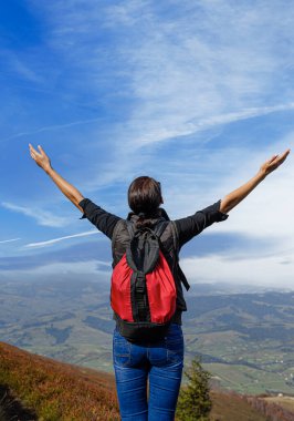 Kırmızı sırt çantalı muzaffer bir kadın yürüyüşçü bir dağın tepesinde duruyor, kolları uçsuz bucaksız mavi gökyüzüne doğru yükseliyor ve aşağıdaki vadiyi süpürüyor..