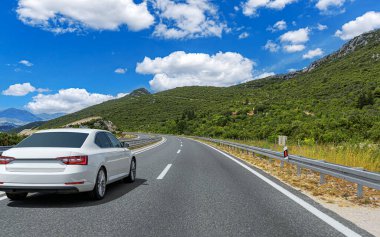 Beyaz bir sedan, sarmal bir asfalt yolda, yemyeşil tepelerin arasından geçiyor. Parlak mavi bir gökyüzünün altında, beyaz bulutların altında..