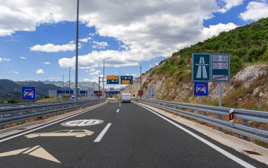 Karavanı olan bir araba otoban gişesine yaklaşıyor ve beyaz bulutlu mavi bir gökyüzünün altında dağlık bir araziye yerleşmiş sınır geçişi yapıyor..