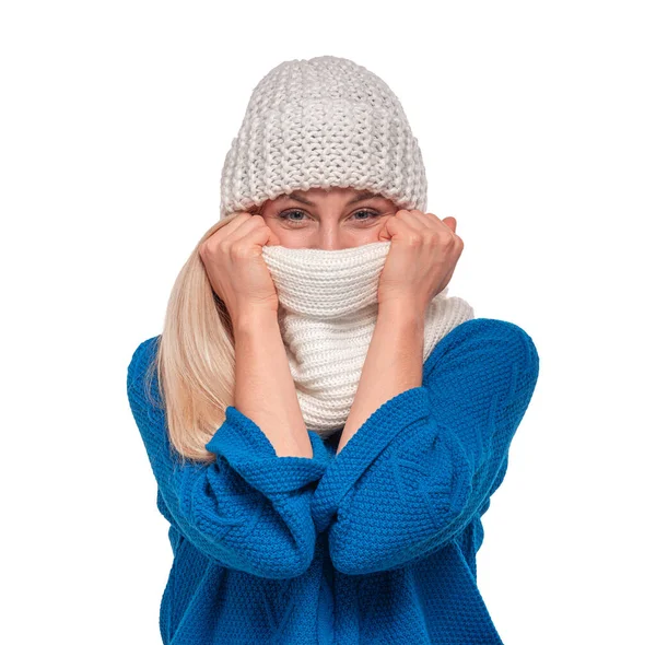 Mavi kazaklı sarışın bir kadın, yüzünün alt kısmını beyaz bir arkaplana karşı aynı bereyi takarken kalın beyaz bir eşarbın arkasına saklar..