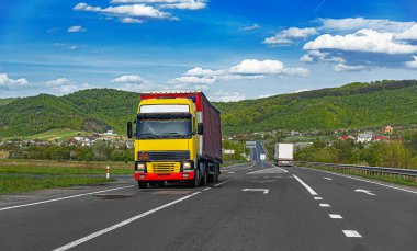 Kırmızı römorklu büyük sarı bir yarı kamyon, beyaz bulutlu mavi bir gökyüzünün altındaki manzaralı dağ manzarası boyunca modern asfalt bir otoyol boyunca ilerliyor..