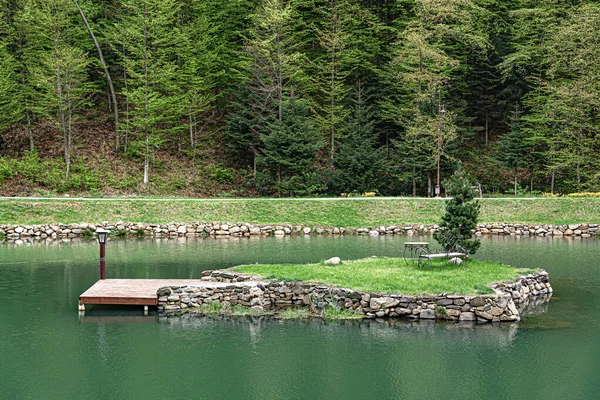 Küçük, insan yapımı bir taş ada, ahşap bir rıhtım ve bir park bankı olan sakin yeşil bir gölün ortasında, yemyeşil bir ormanla çevrili..