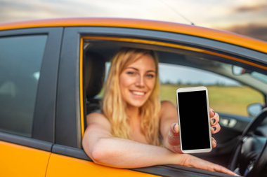 Gülümseyen sarışın bir kadın turuncu bir arabada oturur ve elinde beyaz bir akıllı telefon ile kameraya doğru siyah bir ekran tutar..