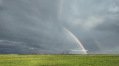 Şiddetli gri bulutların altında yağmur fırtınasından sonra yeşil bir çayırın üzerinde çift gökkuşağı oluştuğunu gösteren hızlandırılmış video. Canlı renkleri, dramatik havayı ve huzurlu kırsal atmosferi yansıtan doğal bir manzara..