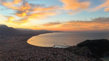 Türkiye 'nin Alanya kentinin geniş bir hava gündoğumu manzarası, şehri, körfezi ve kıyı şeridini sabahın erken saatlerinde sıcak ışıkla aydınlatıyor. Sakin deniz, uzak dağlar ve altın gökyüzü