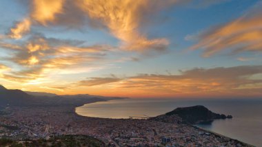 Türkiye 'nin Alanya kentinin geniş bir hava gündoğumu manzarası, şehri, körfezi ve kıyı şeridini sabahın erken saatlerinde sıcak ışıkla aydınlatıyor. Sakin deniz, uzak dağlar ve altın gökyüzü