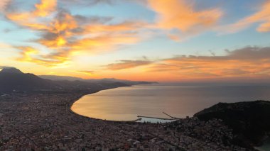 Türkiye 'nin Alanya kentinin geniş bir hava gündoğumu manzarası, şehri, körfezi ve kıyı şeridini sabahın erken saatlerinde sıcak ışıkla aydınlatıyor. Sakin deniz, uzak dağlar ve altın gökyüzü